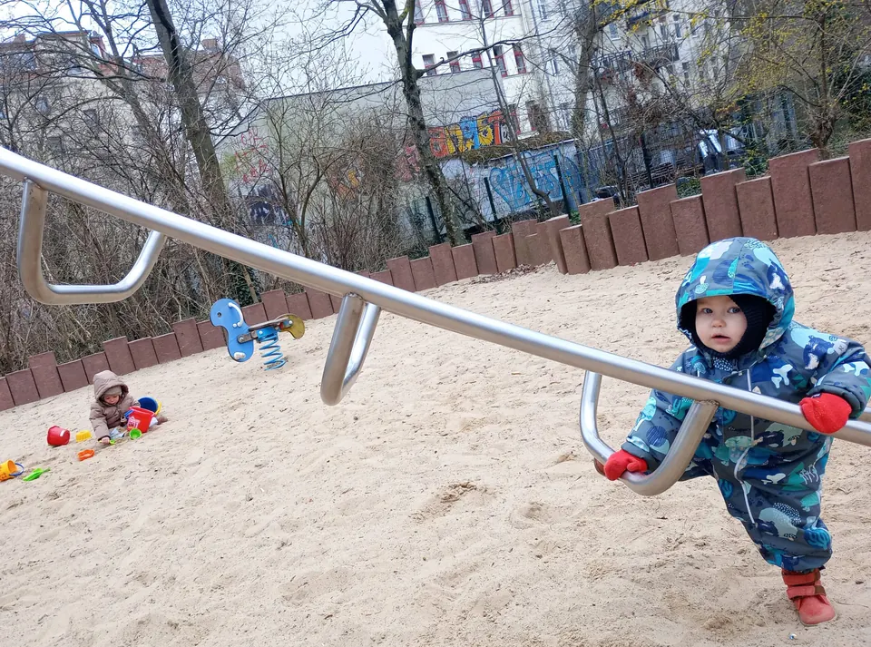 Ein Kind in blauem Overall klettert im Sandkasten an einer Stange