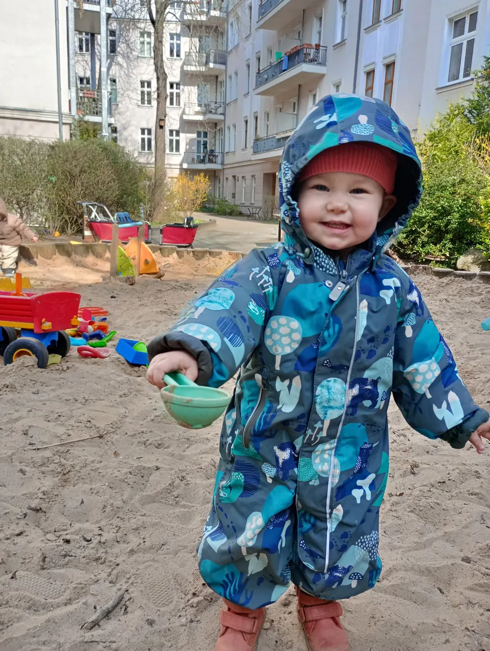 Ein lachendes Kind im Sandkasten hält eine grüne Schale