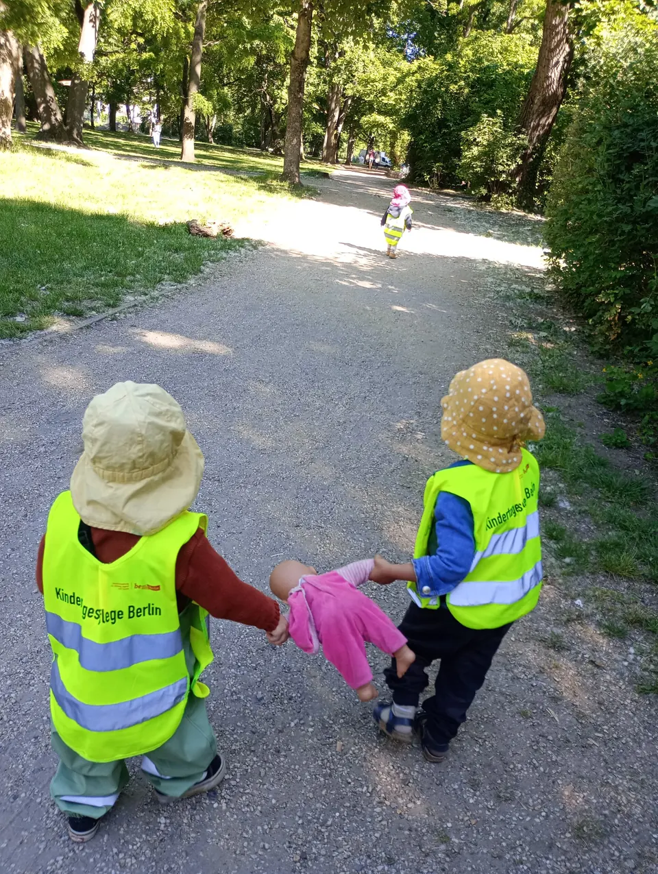 Zwei Kinder in Warnwesten gehen mit einer Puppe durch den Park